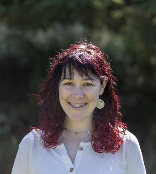 Charis is standing in front of trees smiling and wearing a while blouse.