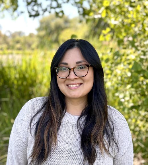 Erika is standing outside in front of trees wearing a white long sleeve shirt and smiling at the camera.
