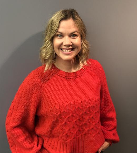 Diana Malarchik is standing in front of a dark brown wall and she is wearing a red sweater smiling at the camera.