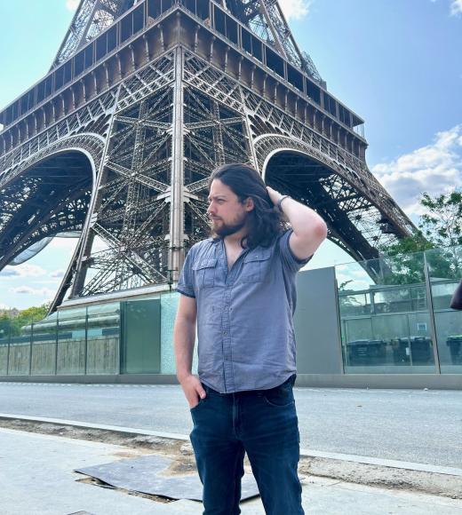John Paulus is wearing a blue shirt and black pants while standing in front of the Eiffel tower.