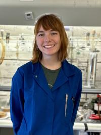 Anna Vernier is standing in a lab with a blue lab coat on and smiling. 