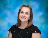 Nicole Keough is smiling in front of a blue background wearing a polkadot shirt.