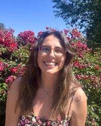 Ece is smiling at the camera in front of the blue sky and red flowers.