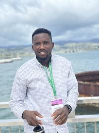 Emmanuel Momoh is standing in front of water wearing a button up long sleeved shirt.