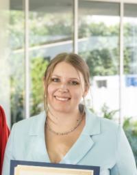 Sabrina is wearing a light blue blazer and smiling for the camera in front of a glass wall while holding an award.