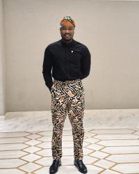 Ibrahim is standing looking at the camera wearing a orange, black, and white pants with a black shirt. He is also wearing a matching kufi.