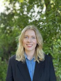Joanna is wearing a sky blue shirt with a navy blazer while smiling at the camera outside.