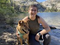 Sage is smiling while kneeling down in front of water with her dog.