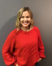 Diana Malarchik is standing in front of a dark brown wall and she is wearing a red sweater smiling at the camera.