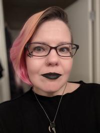 Ste is smiling for the camera with black lipstick and a black shirt while inside a room.