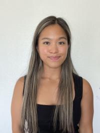 Angel is wearing a black shirt in front of a white wall while smiling for the camera.