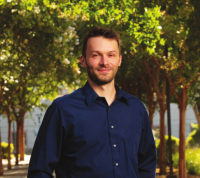 Colton is stadning in front of trees outside while wearing a blue button up long sleeve shirt.